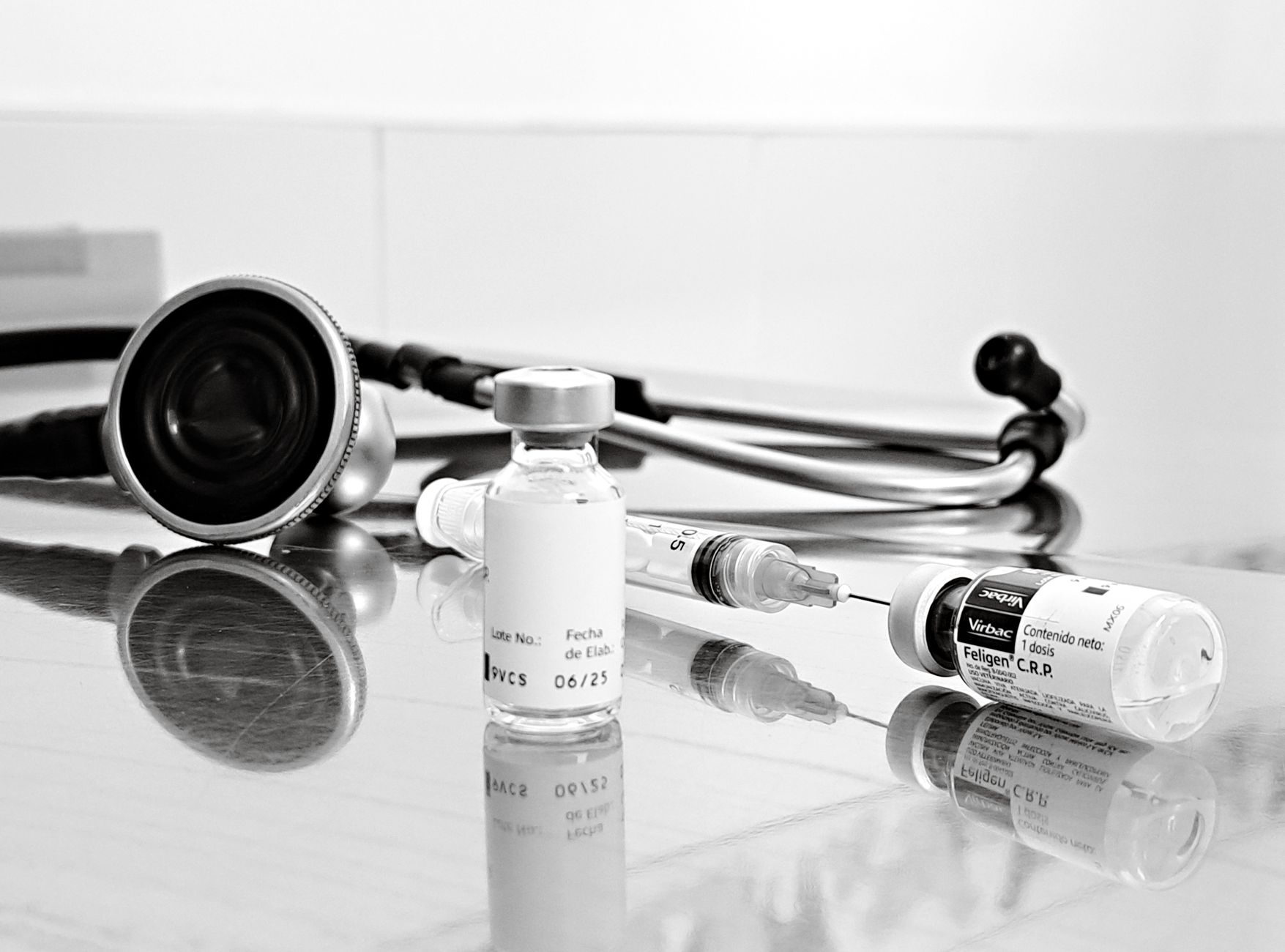 Black and white image of medical stethoscope next to vials and syringe on reflective surface.